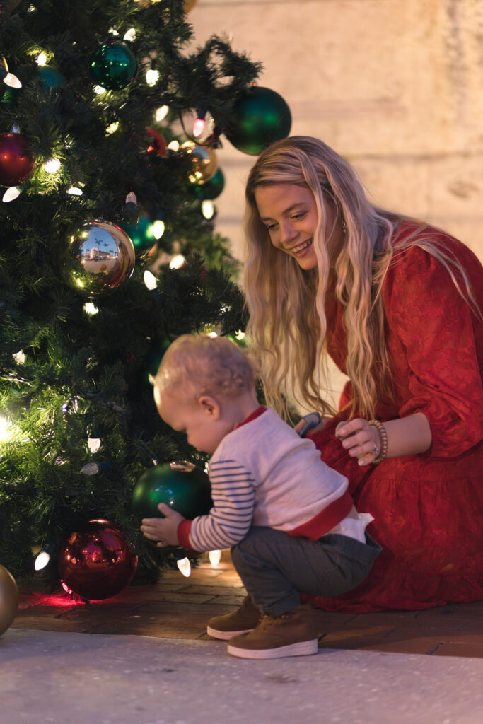 Young boy looks at a large ornament on the Fort Myers Christmas tree, his mom sits behind him and smiles
