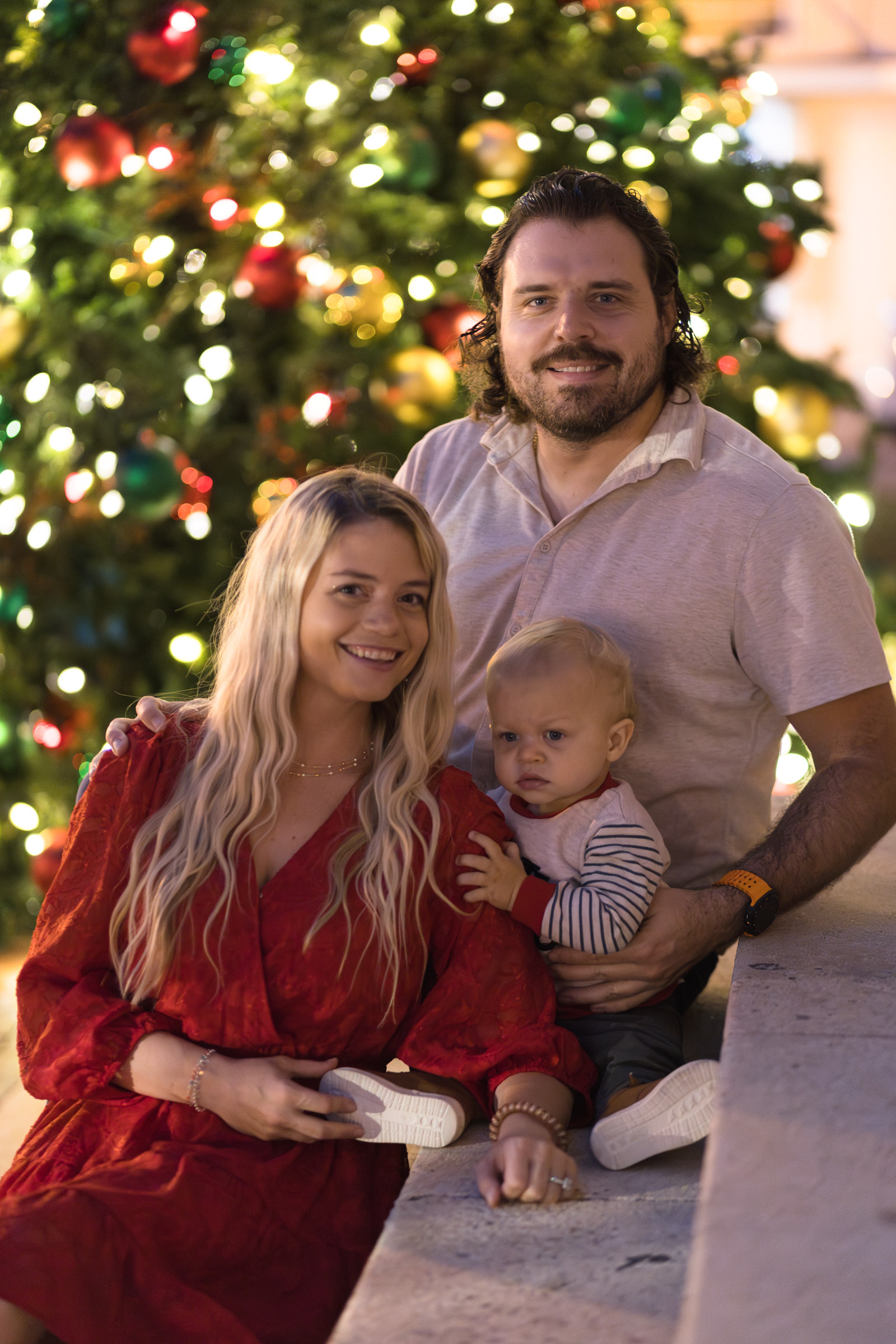 Family of 3 poses on the staircase with a large Christmas tree in the background in Downtown Fort Myers