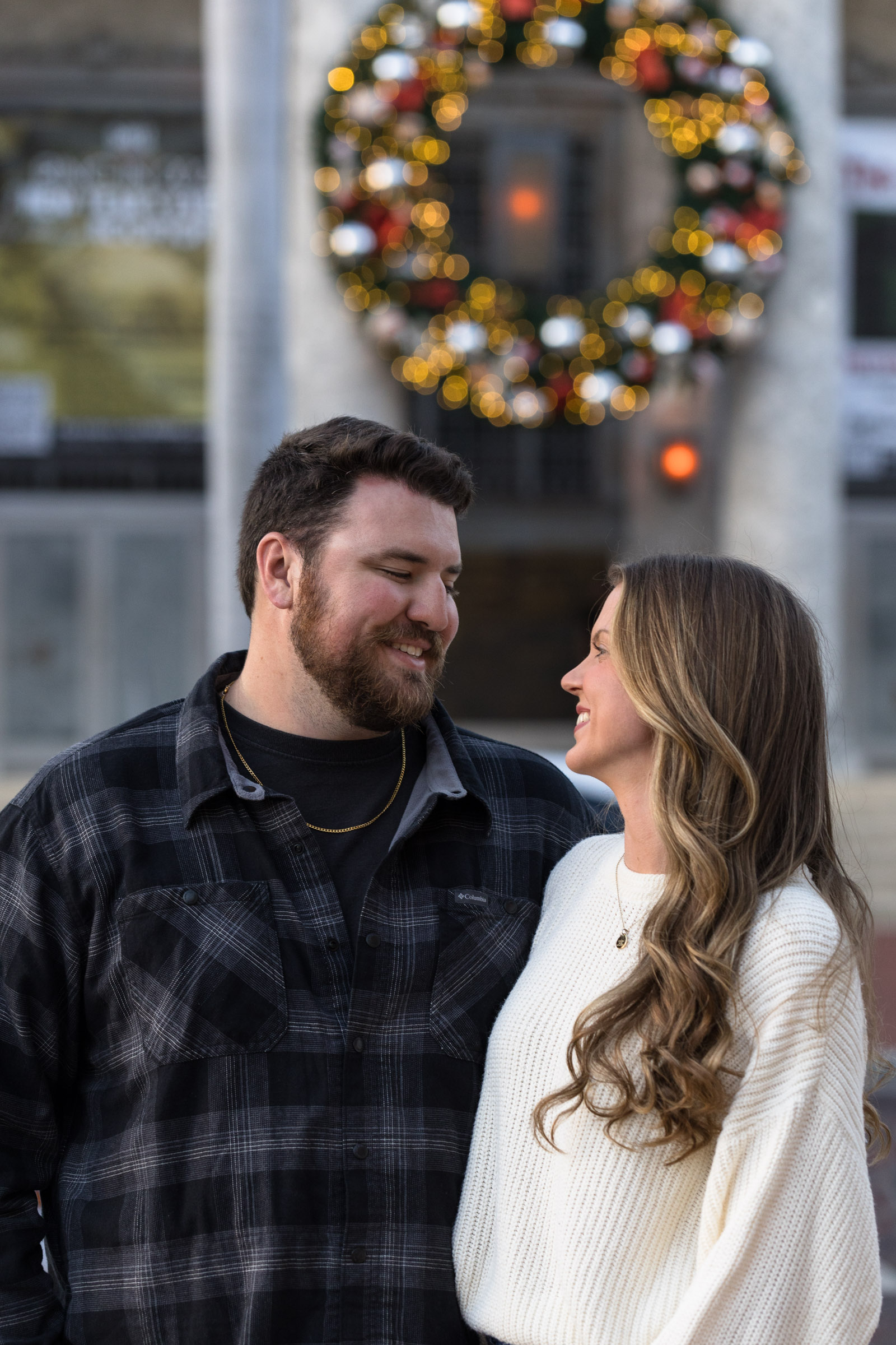 Couple smiles at each other with a blurry Christmas wreath in the background above them in Downtown Fort Myers