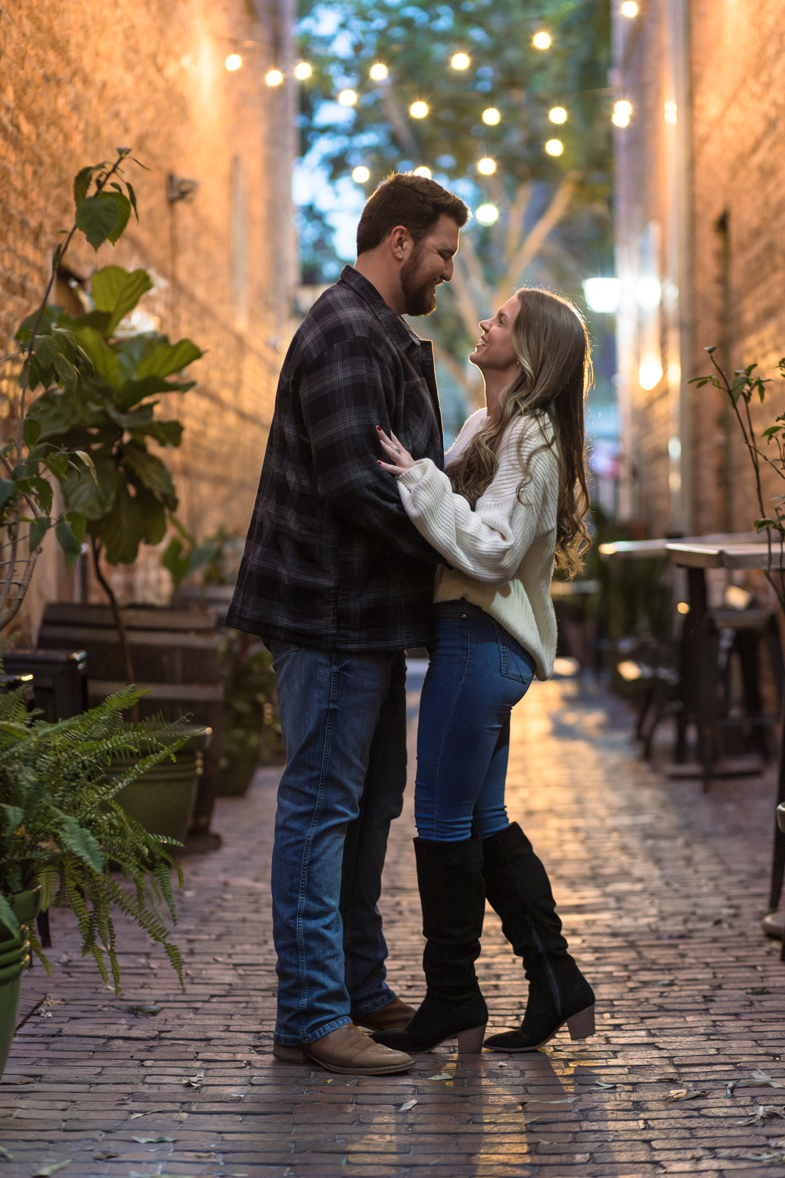 Couple smiles at each other in a cute alley with Christmas lights