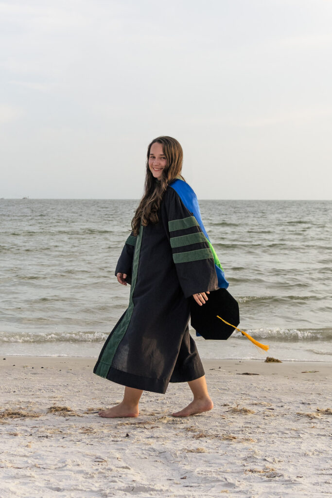 FGCU grad walking in graduation gown and swinging grad cap at the beach