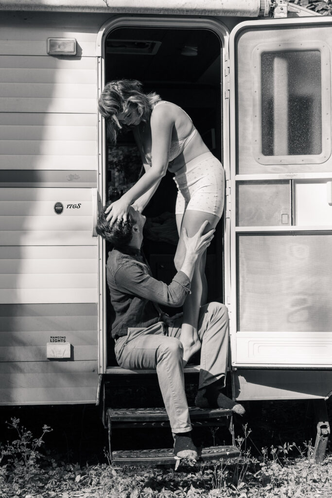 Groom sits on the step of campervan airbnb, his bride takes a moment during getting ready to play with his hair