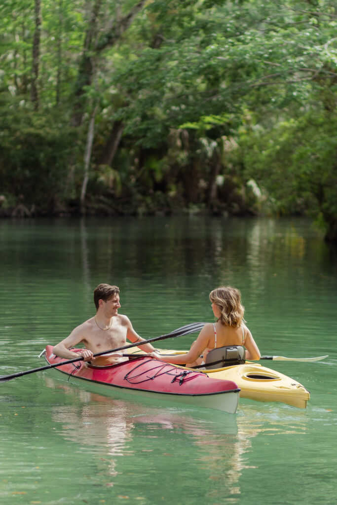 Couple in two separate kayaks in a clear river under a canopy of trees during their adventure kayaking photoshoot