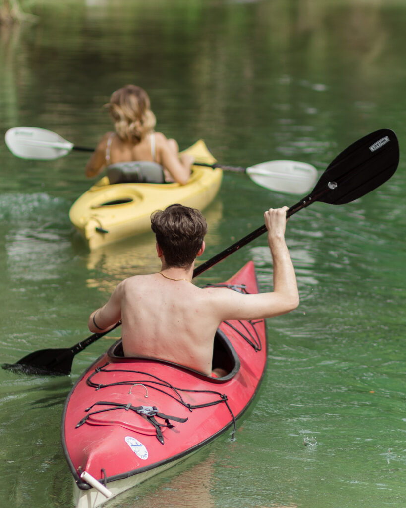 Man paddles in a red kayak behind his wife in a yellow kayak at their kayaking photoshoot