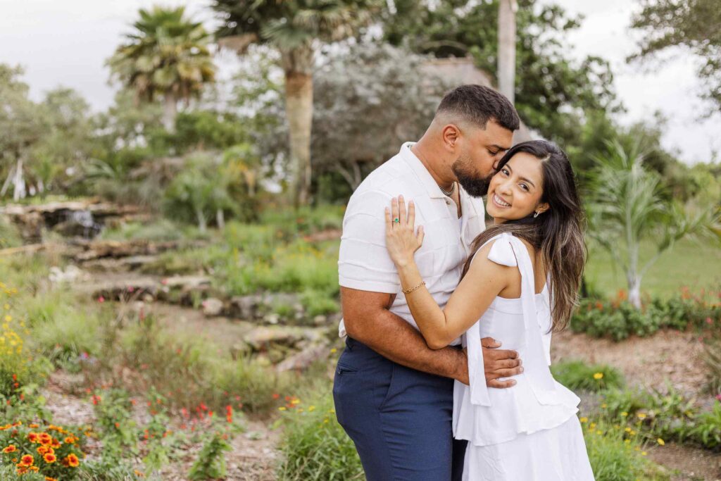 Man kisses fiancée's cheek as she smiles at the camera in front of a stream and flowers.
