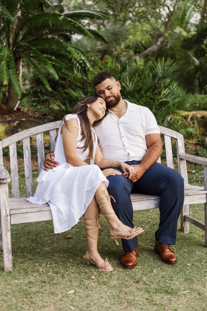 Woman rests her head on fiancé's shoulder on a Naples Botanical Garden bench