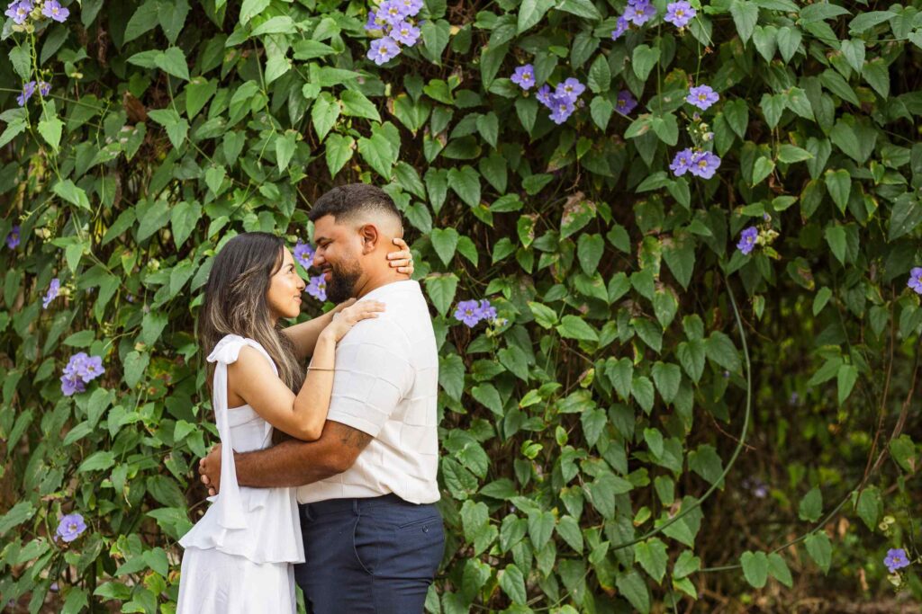 Couple looking at each other and smiling in front of a wall of leaves and purple flowers