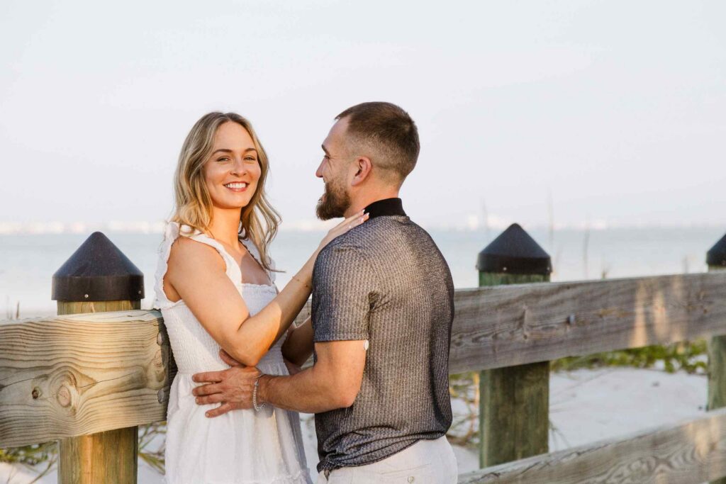 Woman leans against fence and smiles at the camera, her fiancé holds her wait and smiles at her
