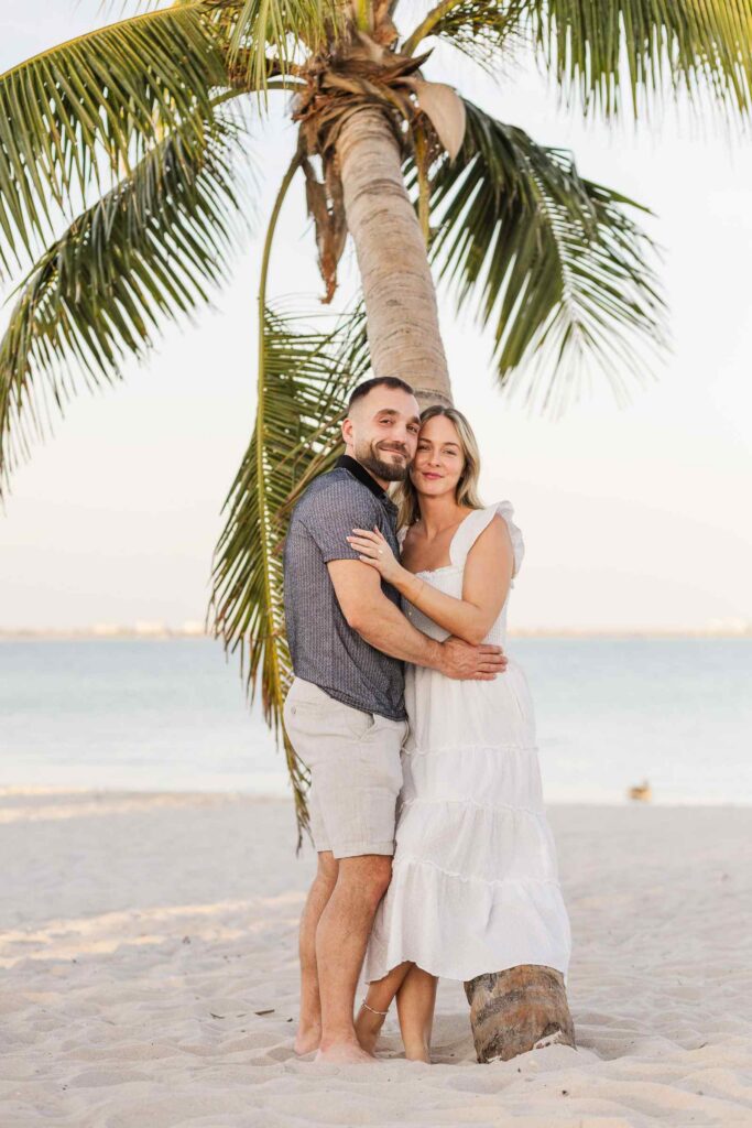 Couple smiles at the camera while leaning against a palm tree