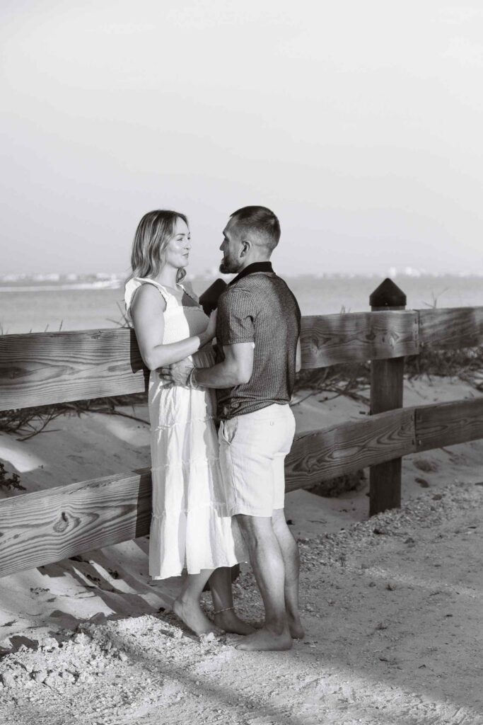 Black and white image, woman leaning against fence with her fiancé in front of her holding her waist