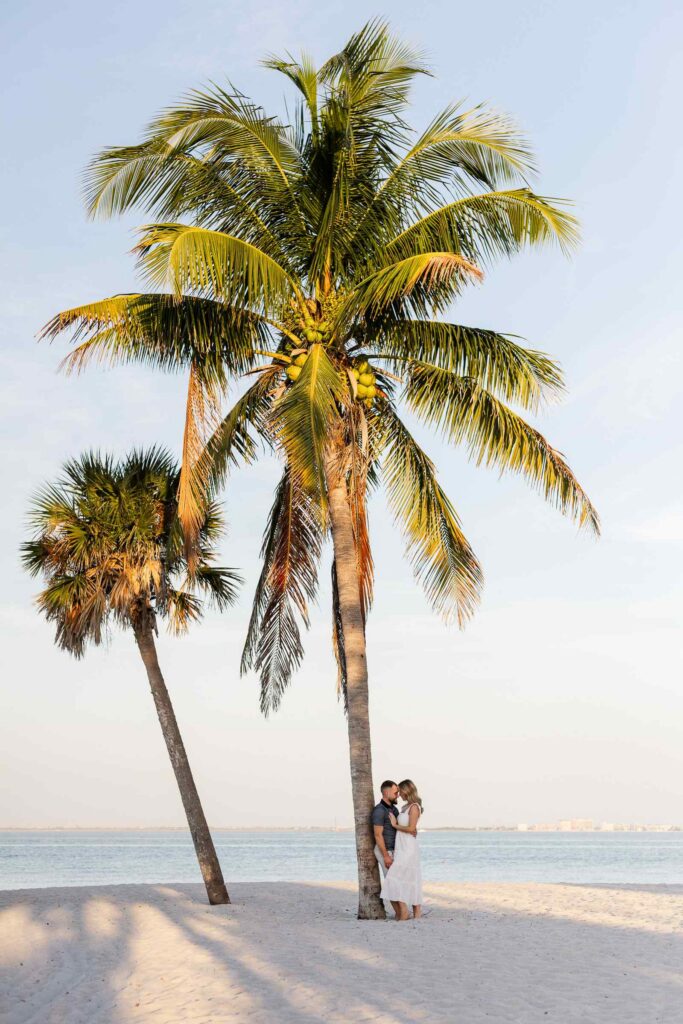 A large palm in the middle with a smaller one to the left, a couple is small in the frame leaning against the base of the large palm tree