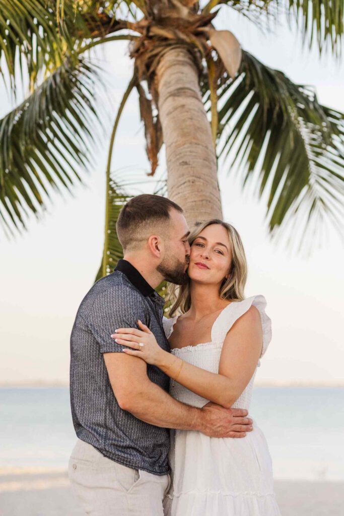 Man kisses fiancée's cheek as she smiles at the camera, the top of a palm tree is behind them above their heads
