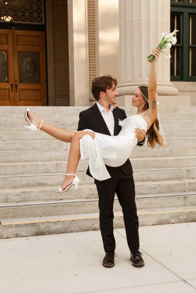 Groom carries bride as she lifts her bouquet up after their Fort Myers Courthouse wedding
