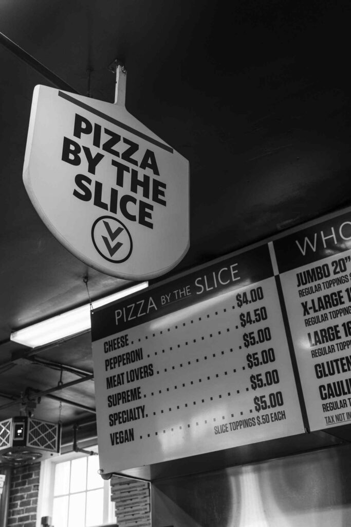 Greyscale "pizza by the slice sign" by the menu at Downtown House of Pizza