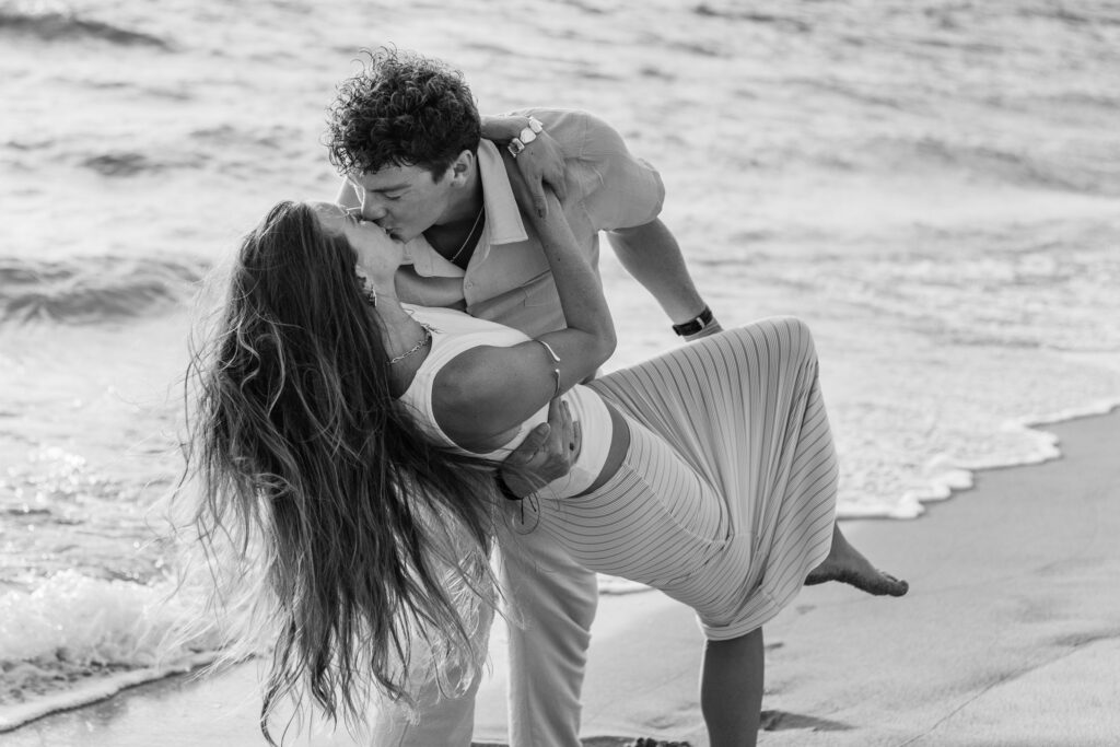 Black and white image of man dipping fiancee for a kiss on the shoreline
