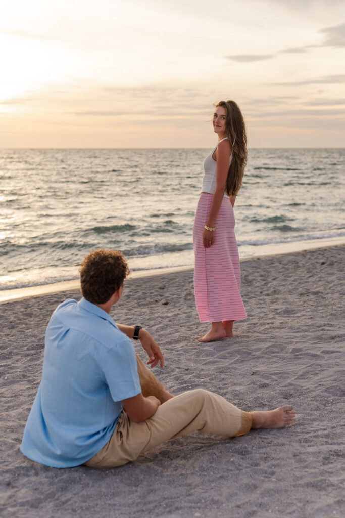 Man sits on the beach and looks at his fiancee standing in front of him