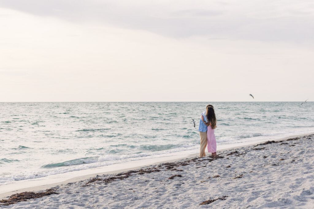 Woman and boyfriend hug in a wide shot of the beach with 3 seagulls flying near them