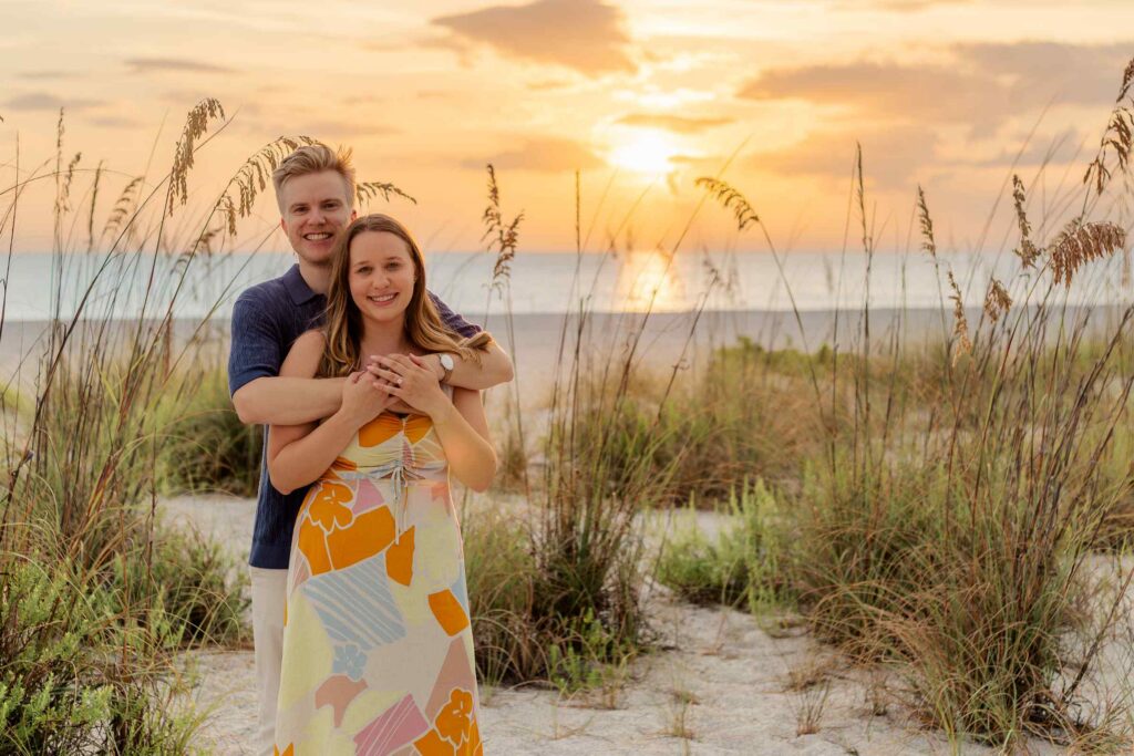 Couple posing in the middle of beach grass with a bright orange sunset and clear blue ocean water behind them
