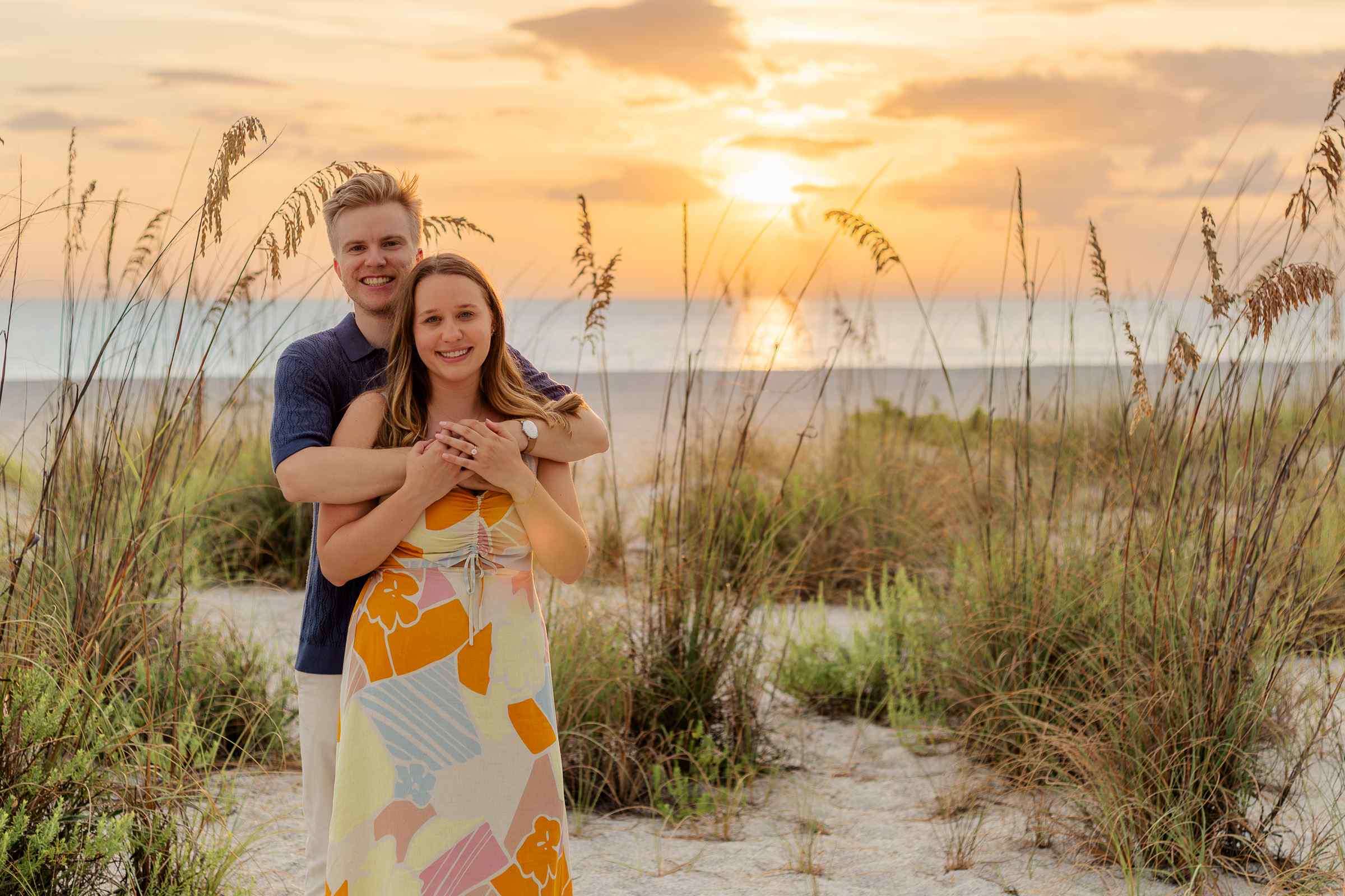Couple posing in the middle of beach grass with a bright orange sunset and clear blue ocean water behind them