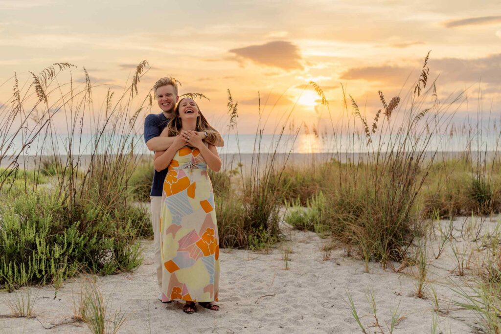 Couple laughing in the middle of beach grass with a bright orange sunset and clear blue ocean water behind them