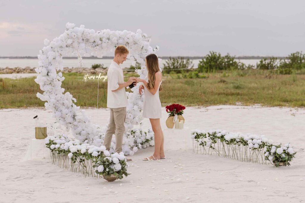 Couple in a heart arch setup on the beach, the man is placing a ring on the woman's finger