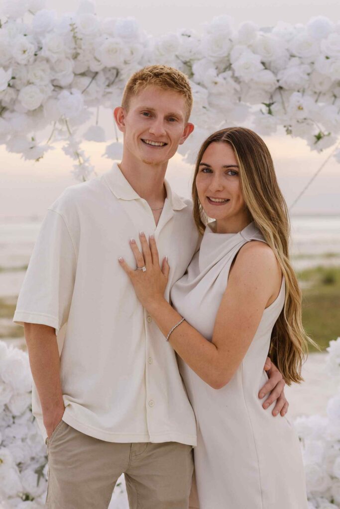 Couple poses and smiles at the camera in the middle of a white heart arch