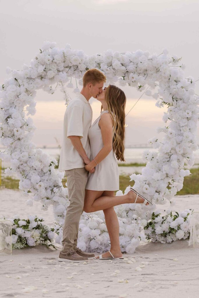 Couple kisses and woman pops her foot up in the middle of a white heart arch