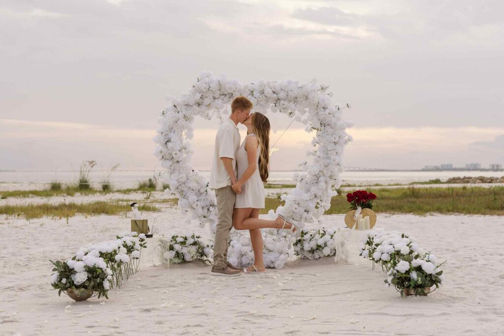 Couple kisses and woman pops her foot up in the middle of a white heart arch
