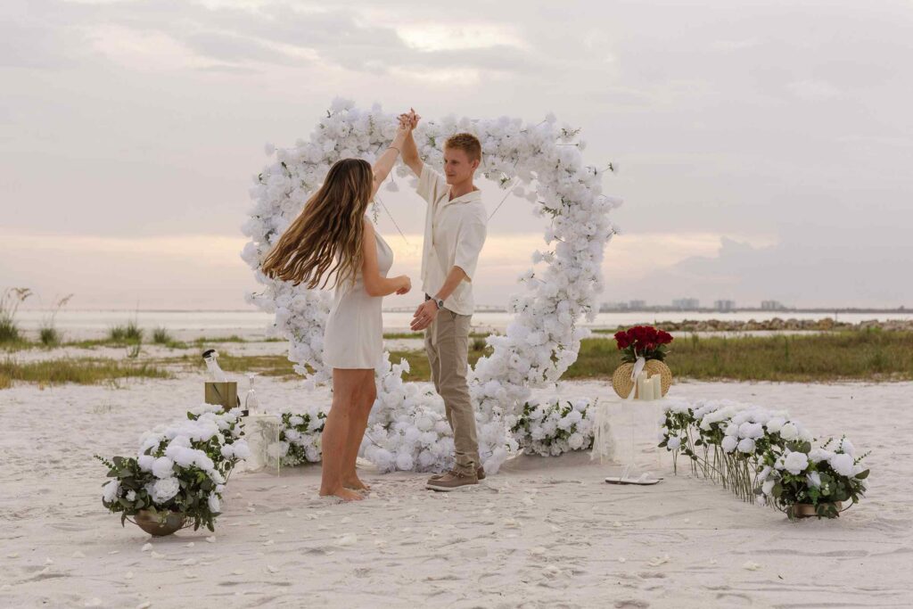 Couples dances and man twirls woman in the middle of a heart arch proposal setup